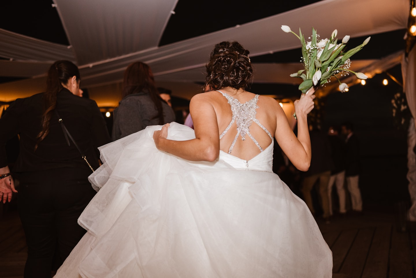 Novia con su vestido y flores caminando bajo luces de guirnalda.