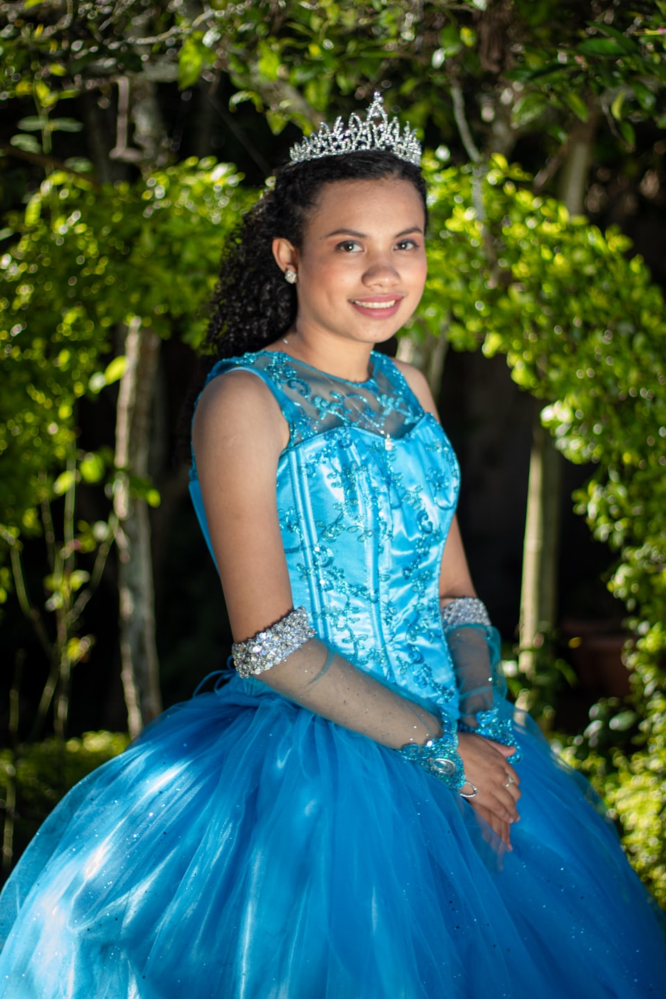 Joven sonriente con vestido azul de quinceañera y tiara al aire libre.